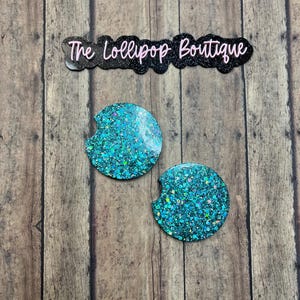 May include: Two teal glitter car coasters with a crescent-shaped cutout, set against a wooden background. The coasters are round and feature a sparkling, textured surface. The text "The Lollipop Boutique" is above the coasters.