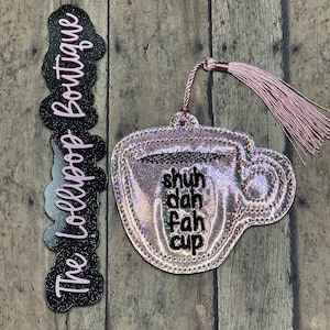 May include: A pink and silver glittery coffee cup-shaped ornament with the words "shuh dah fah cup" embroidered on it. It has a pink tassel and a black and pink glittery tag that says "The Lollipop Boutique".