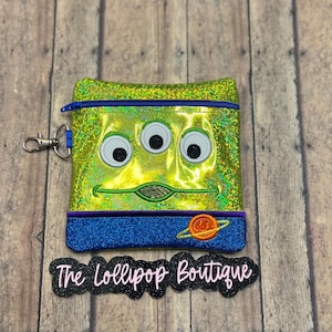May include: A green, glittery pouch featuring the face of a three-eyed alien with a blue zipper and trim. The pouch has a blue and purple bottom section with an orange planet design. Includes a silver clip. The text "The Lollipop Boutique" is below.