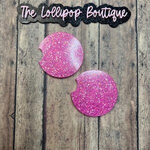 May include: Two pink glitter car coasters with a semi-circle cutout. The coasters are round and have a shiny, textured surface. The text "The Lollipop Boutique" is above the coasters.