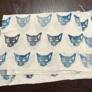 May include: A stack of fabric squares with a repeating pattern of blue cat faces printed on a white background.