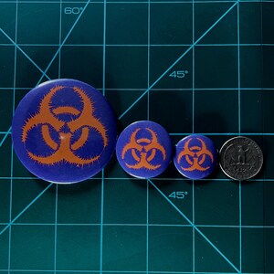 May include: Three biohazard symbol buttons on a green cutting mat. The largest button is blue with an orange biohazard symbol. The two smaller buttons are blue with orange biohazard symbols. A silver US quarter is included for size comparison.