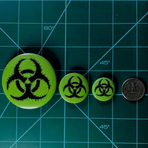 May include: Three green and black biohazard symbol buttons of different sizes, next to a silver US quarter.