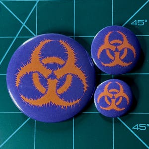 May include: Three biohazard symbol buttons in different sizes. The largest button is purple with an orange biohazard symbol. The medium button is purple with an orange biohazard symbol. The smallest button is purple with an orange biohazard symbol.
