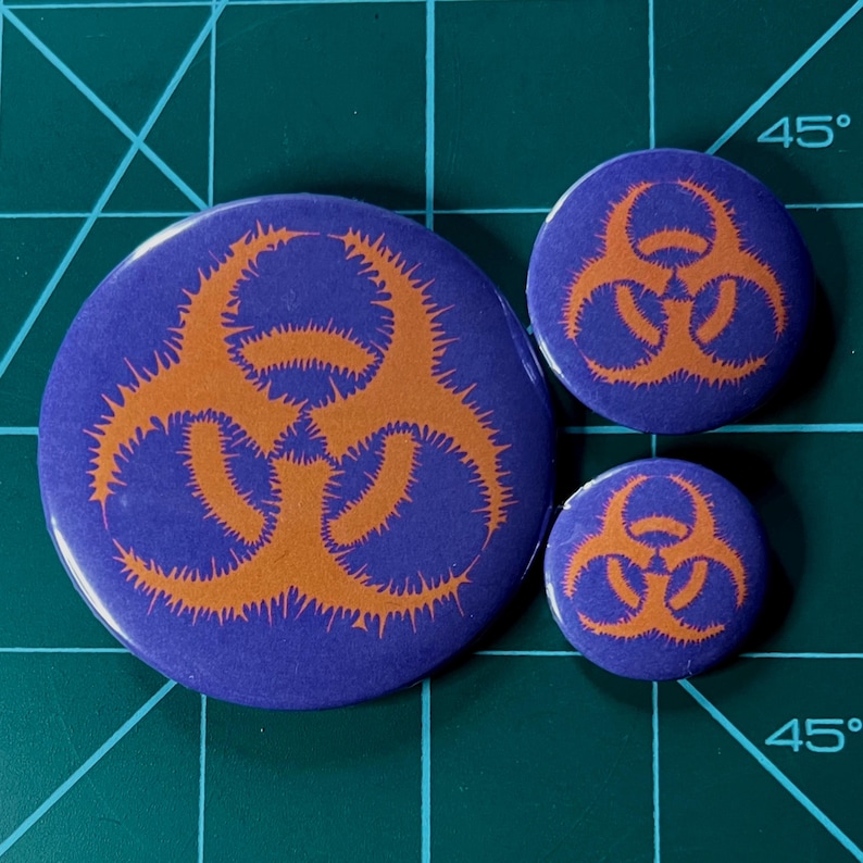 May include: Three biohazard symbol buttons in different sizes. The largest button is purple with an orange biohazard symbol. The medium button is purple with an orange biohazard symbol. The smallest button is purple with an orange biohazard symbol.