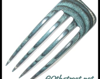 Puede incluir: Una horquilla para el cabello de cuatro puntas con un patrón de vetas de madera en color verde azulado y gris oscuro. El accesorio presenta un borde superior curvo y puntas afiladas. El texto 60thstreet.net aparece en la esquina inferior.
