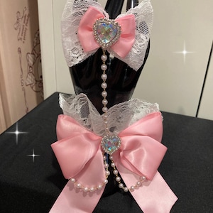 May include: Two pink satin bows with white lace trim and a heart-shaped rhinestone pendant. Each bow is adorned with a string of pearls. The bows are displayed on a black mannequin hand.