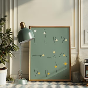 May include: A framed art print with a gold border features minimalist line art on a muted green background. The artwork includes black lines and yellow and white circular accents. A teal desk lamp with a gold top is in the foreground.