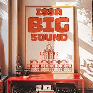 Könnte beinhalten: Ein gerahmtes Poster mit den Worten "ISSA BIG SOUND" in orangefarbenen Blockbuchstaben. Darunter befindet sich eine Grafik einer Person, die über einem Stapel von Lautsprechern meditiert. Das Poster wird auf einem roten Tisch ausgestellt.