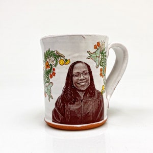 Könnte beinhalten: Weiße Keramiktasse mit einem braunen Bild einer Person mit Brille und einem dunklen Gewand. Die Tasse ist mit grünen, orangefarbenen und gelben Blumendesigns verziert.