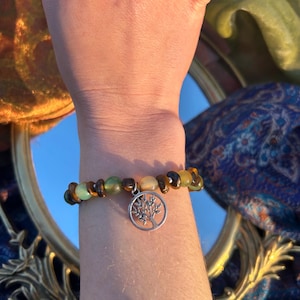 May include: A beaded bracelet with a silver tree of life charm. The beads are brown, green, and yellow.