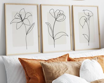 May include: Three framed botanical art prints. Each print features a black line drawing of a different flower against a neutral background. The frames are light wood. The prints are displayed above a bed with orange, brown, and white pillows.