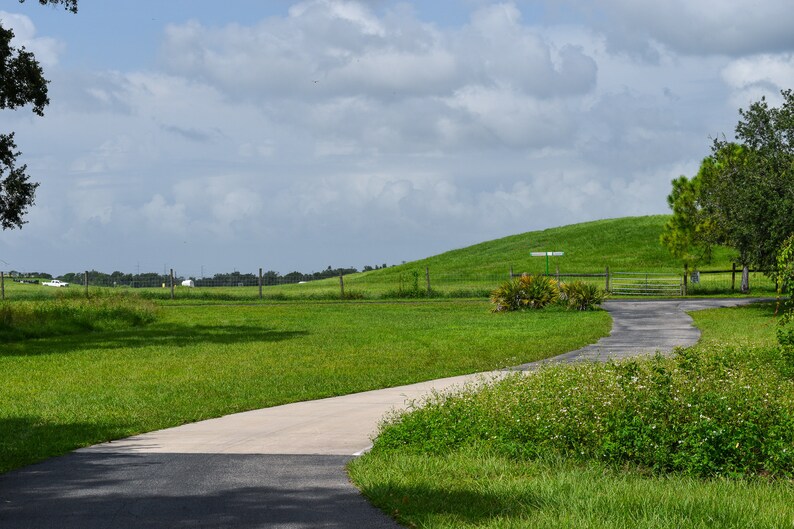 May include: A winding paved driveway leads to a grassy field with a rolling green hill in the background. The sky is partly cloudy.