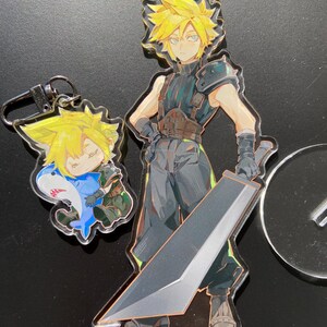 May include: Acrylic charms featuring a character with spiky yellow hair. One charm depicts the character with a large sword, while another shows a smaller version in a shark costume. The charms are clear with colorful illustrations.