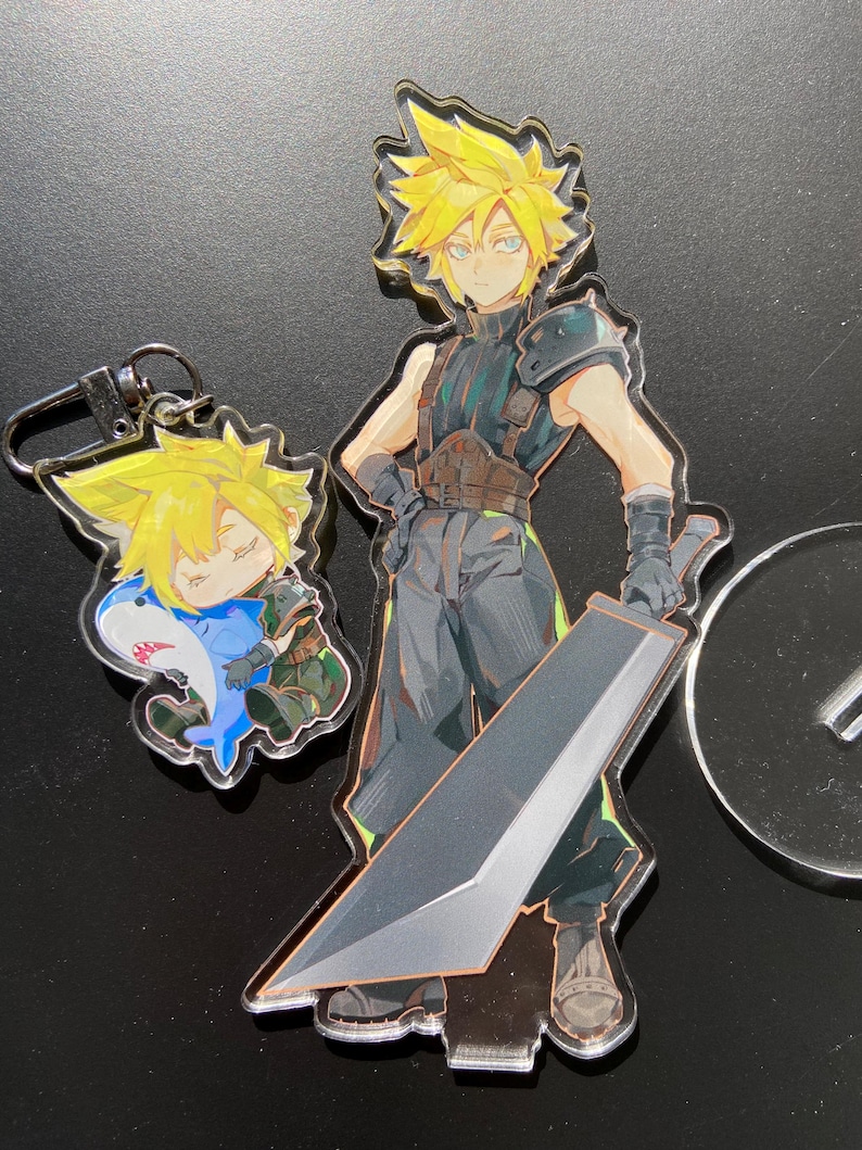 May include: Acrylic charms featuring a character with spiky yellow hair. One charm depicts the character with a large sword, while another shows a smaller version in a shark costume. The charms are clear with colorful illustrations.
