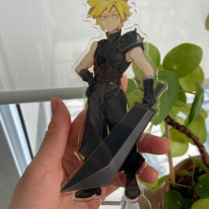 May include: A clear acrylic figure of a character with spiky yellow hair, wearing a black outfit and holding a large black sword. The figure is standing on a clear base. The character has a determined expression.