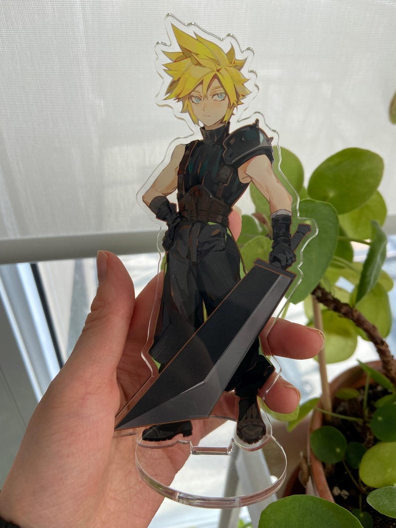 May include: A clear acrylic figure of a character with spiky yellow hair, wearing a black outfit and holding a large black sword. The figure is standing on a clear base. The character has a determined expression.