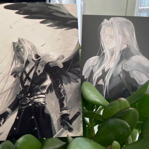 May include: Two art prints featuring a character with long silver hair and armor. One print shows the character with a wing and a sword, the other shows the character in profile. The prints are set against a black and white background.