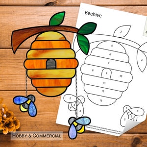 May include: A stained glass beehive pattern with numbered pieces for a craft project. The beehive is yellow and orange with a small entrance hole. Two bees are hanging from the bottom of the beehive.