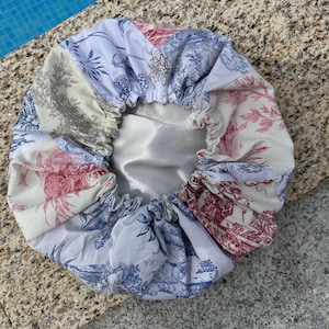 May include: A white satin lined shower cap with a patchwork design featuring a variety of floral and nature-inspired patterns in shades of blue, red, and gray.