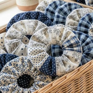 May include: A wicker basket filled with handmade fabric coasters. The coasters feature various blue and white patterns, including polka dots, floral prints, and plaid designs. The coasters are circular and appear to be padded.