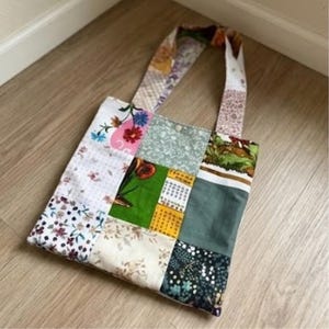 Può includere: Una borsa tote patchwork realizzata con vari quadrati di tessuto caratterizzati da motivi diversi, tra cui stampe floreali, verde a tinta unita e illustrazioni botaniche. La borsa include due manici in tessuto abbinati e un design quadrato.