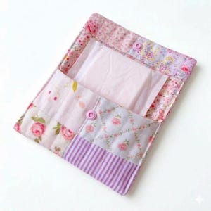 Pode incluir: Uma bolsa de tecido acolchoado em tons de rosa, lavanda e branco, com padrões florais e listrados. A bolsa está aberta, revelando um penso higiénico. Dois fechos de pressão rosa prendem a bolsa.