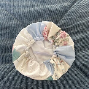 Pode incluir: Touca de patchwork com secções de tecido floral, de bolinhas e liso em tons pastel. A touca tem uma borda elástica franzida e um interior de cetim branco. Os tecidos incluem azul claro, rosa e creme.