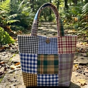 Pode incluir: Uma sacola tote patchwork com uma variedade de padrões xadrez e vichy em azul, preto, marrom e verde. A sacola tem um fecho de botão de madeira e uma alça resistente. A sacola está em um caminho de pedra.