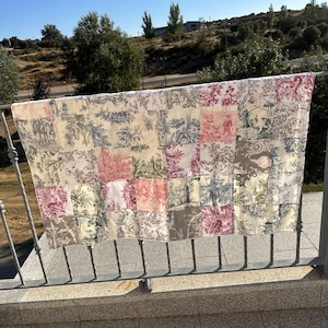 May include: A patchwork quilt featuring toile patterns in cream, red, grey, and blue. The quilt is displayed over a metal railing outside, highlighting the different fabric squares and designs.