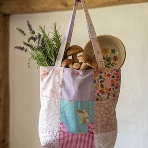 Puede incluir: Un bolso tote patchwork con una variedad de telas florales y estampadas, colgado de un gancho. La bolsa está llena de hierbas frescas, setas y un cuenco decorativo con un diseño floral. Los colores de la bolsa incluyen rosa, azul y lavanda.