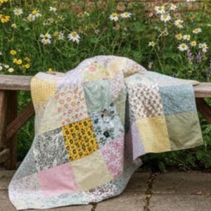 Op de afbeelding: Een patchwork quilt in pastelkleuren, gedrapeerd over een houten bank. De quilt is gemaakt van verschillende stoffen met patronen, waaronder bloemen- en geometrische ontwerpen. De achtergrond toont een tuin met madeliefjes.