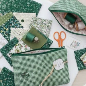 May include: A collection of green and white patterned fabrics, sewing accessories, and two zipped pouches. The pouches and fabric have floral and gingham designs. Orange scissors, thread spools, and a thimble are also visible.