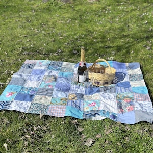 May include: A blue and white patchwork picnic blanket with a bottle of champagne and a glass on top of it. A wicker basket is also on the blanket.