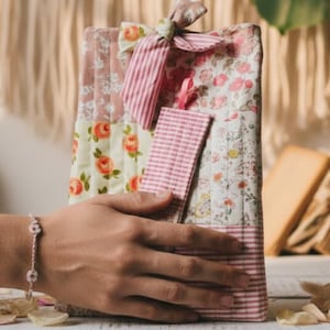 Puede incluir: Una funda de libro hecha a mano con estampados florales y a rayas en rosa, blanco y melocotón. La funda está atada con un lazo a juego e incluye un marcapáginas a rayas rosas. La estética general es vintage y femenina.