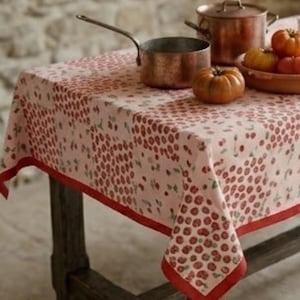 May include: A rectangular tablecloth with a red cherry pattern on a light pink background. The tablecloth has a red border and is draped over a dark wooden table. Copper pots and a bowl of tomatoes are on the table.