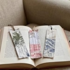 Pode incluir: Três marcadores de tecido repousam nas páginas abertas de um livro. Os marcadores apresentam designs exclusivos: um com uma estampa botânica verde e preta, outro com um padrão de patchwork vermelho e branco e um terceiro com linhas abstratas azuis e brancas. Cada um tem um cordão.