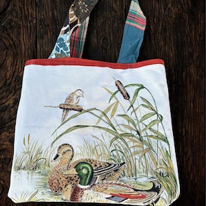 Könnte beinhalten: Eine weiße Tragetasche mit einer Naturszene mit Enten, einem Vogel und Schilf. Die Tasche hat eine rote Einfassung und gemusterte Griffe in verschiedenen Farben. Das Kunstwerk zeigt einen Teich mit Enten und einem Vogel, der auf einem Schilf sitzt.