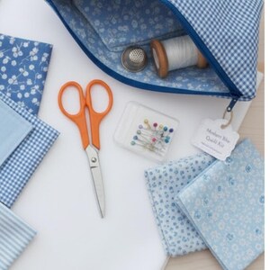 Pode incluir: Uma cole&ccedil;&atilde;o de materiais de costura, incluindo tesouras laranja, alfinetes, quadrados de tecido e uma bobina de linha branca. Uma bolsa com z&iacute;per azul xadrez cont&eacute;m uma dedaleira e mais linha. A etiqueta diz "Modern Blue Quilt Kit."