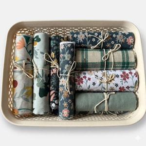 Op de afbeelding: Een beige rechthoekige mand gevuld met opgerolde stoffen doeken. De doeken hebben bloemen- en ruitpatronen in verschillende kleuren, waaronder groen, roze en wit. Elke doek is vastgebonden met touw. De mand staat tegen een witte achtergrond.