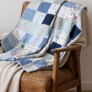 Könnte beinhalten: Eine Patchwork-Decke bestehend aus verschiedenen quadratischen Stoffstücken in Blau-, Weiß- und Cremetönen. Die Decke zeigt diverse Muster, darunter einfarbiges Marineblau, hellblaues Vichy-Karo, florale Drucke und feine vertikale Streifen.