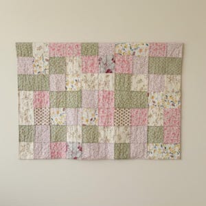 Può includere: Una trapunta patchwork con una varietà di motivi floreali e a pois nei toni del rosa, verde e crema. La trapunta è composta da blocchi quadrati e ha una texture trapuntata.