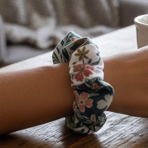 May include: A patterned scrunchie in shades of green, white, and pink, wrapped around a wrist. The scrunchie features a floral design. The fabric appears soft, suggesting it's for hair styling. The scrunchie is a popular hair accessory.