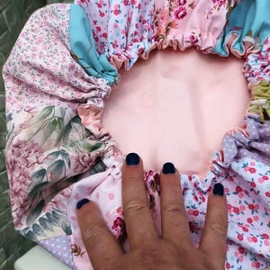 Pode incluir: Uma touca com padrão floral com interior rosa. A touca apresenta uma variedade de tecidos com desenhos florais e de bolinhas em tons de rosa, azul e roxo. As bordas são franzidas, criando um efeito de babado.