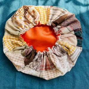 Puede incluir: Un gorro de patchwork con un interior naranja vibrante. El exterior presenta una variedad de patrones de tela, incluyendo diseños florales, cuadros y paisley en tonos beige, amarillo, marrón y verde. El gorro está fruncido en la abertura con un ribete rojo.