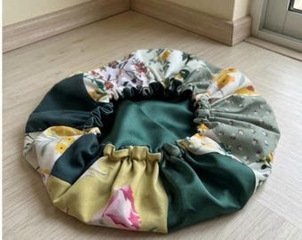 May include: A circular fabric bonnet featuring a patchwork design of solid dark green panels and floral patterned sections in shades of yellow, pink, and white. The accessory has a gathered elastic edge and a smooth, solid dark green interior lining.