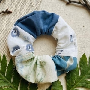 May include: A handmade scrunchie with a patchwork design. The fabric is a mix of blue and white patterns, including floral and geometric prints. The scrunchie is surrounded by green fern leaves and twigs.