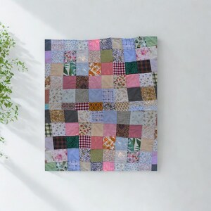 May include: A patchwork quilt with a variety of fabric squares in shades of pink, blue, green, and beige. The quilt is made of many different patterns and textures, creating a unique and colorful design. The quilt is displayed on a white wall.