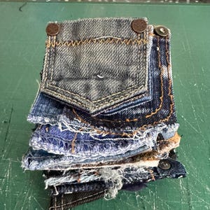 May include: A pile of denim fabric offcuts in shades of blue and grey, with visible stitching and metal button details. The edges are frayed, creating a distressed look. The pieces are likely from jeans or similar garments.