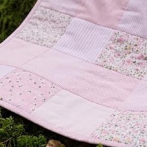 Pode incluir: Uma colcha de retalhos com uma variedade de quadrados de tecido rosa claro. O design inclui quadrados rosa sólidos, padrões de riscas brancas e rosas e estampados florais com pequenas flores rosas. A colcha tem uma borda costurada e textura macia.